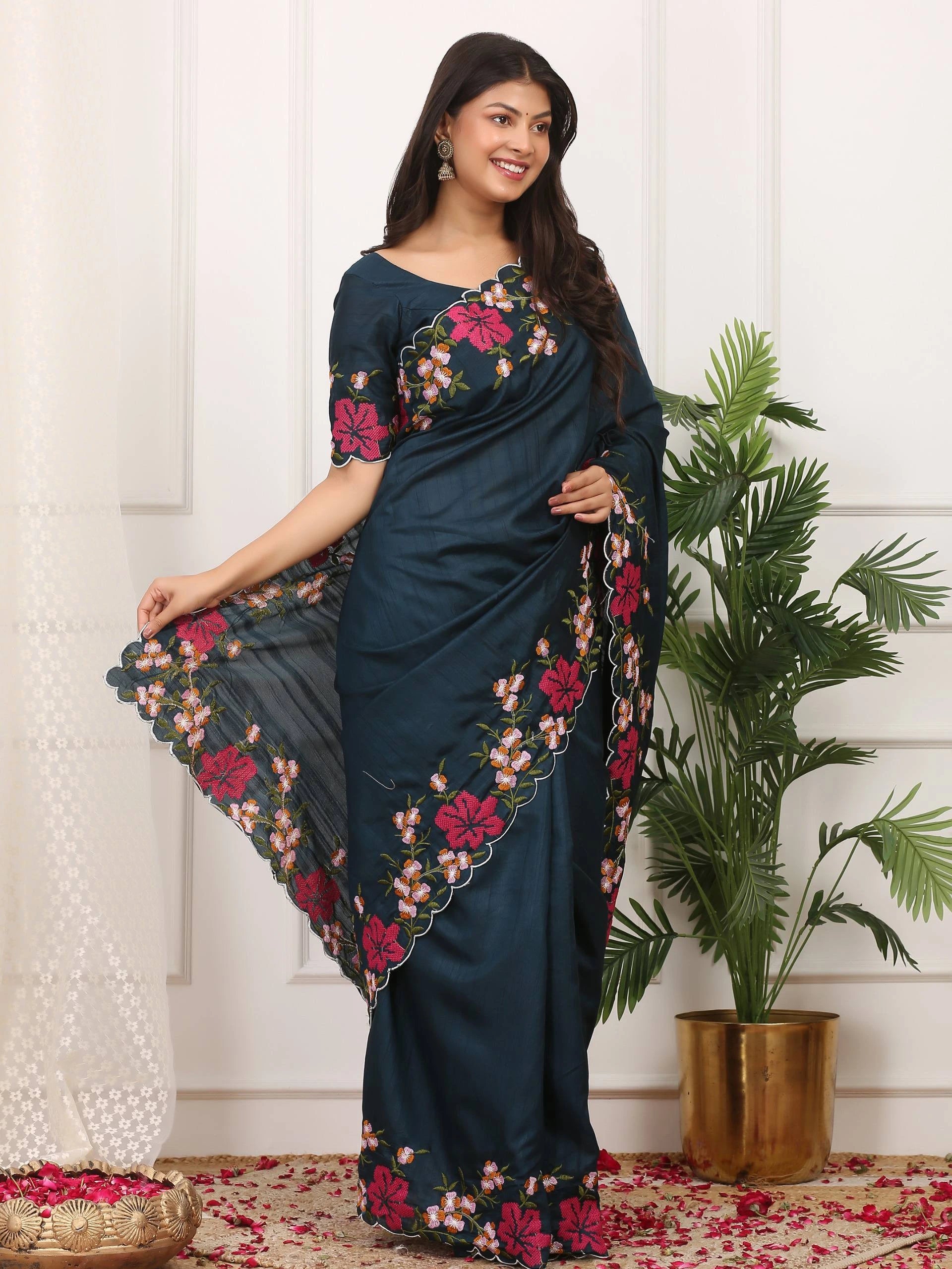 Woman wearing a dark blue saree with floral patterns in a room with a plant and decorative items.